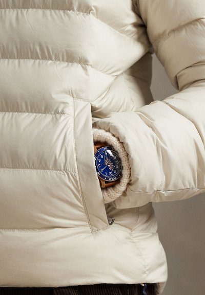 Smėlio spalvos pūkinė striukė su horizontaliu dygsniu; kišenėje iš dalies matomas mėlynas laikrodis su rudu dirželiu, apsuptas megztos pamušalo.