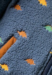 Tumši zils vilnas jaka ar teksturētu virsmu. Iezīmējas daudzkrāsu lapu rakstiem oranžā un zaļā krāsā, ar kontrastējošu brūnu oderi.