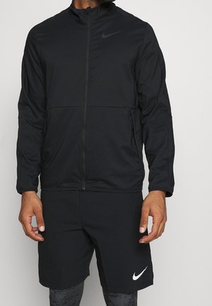 Homme portant une veste noire zippée et un short noir avec des logos Nike blancs, debout devant un fond clair uni.