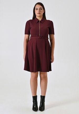 Frau steht in einem burgunderfarbenen Kurzarmkleid mit Gürtel und Reißverschluss vorne, kombiniert mit schwarzen Stiefeletten, vor einem schlichten Hintergrund.
