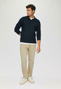 Diepblauwe gebreide pullover met een kraag, crèmekleurige onderlaag, lichtbeige jeans en witte sneakers. Model poseert casual met één hand achter de rug.