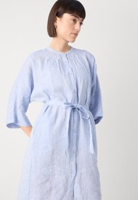Svetlo modra srajčna obleka iz lanu s polovičnimi rokavi, gumbi spredaj in pasom. Ima subtilno teksturo in naborke na izrezu.