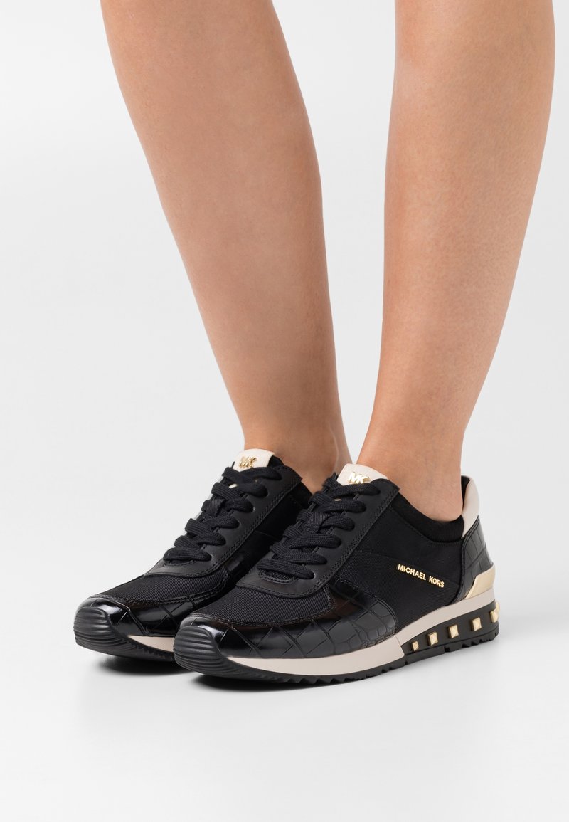 Jambes portant des baskets Michael Kors noires et beiges avec motif matelassé, logo doré et semelle cloutée sur fond blanc uni.