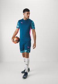 Puma-Sportausrüstung mit einem türkisfarbenen Shirt mit gemusterten Ärmeln, pinken Akzenten, passenden Shorts, weißen Socken und blauen Fußballschuhen. Hält einen Fußball.