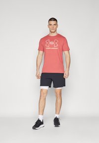 Rød t-skjorte med hvitt "Under Armour"-logo, sammen med svarte shorts. Modellen har på seg svarte joggesko og hvite sokker, og står mot en grå bakgrunn.