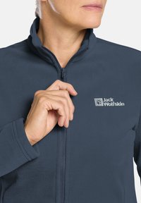 Marineblaue Fleecejacke mit hohem Kragen, Reißverschluss vorne und gesticktem Jack Wolfskin-Logo. Weiche Textur und enganliegendes Design.