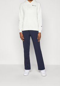 Bílá mikina s kapucí a tmavě modré tepláky. Mikina má přední kapsu a logo na hrudi. Bílé tenisky doplňují celý outfit.