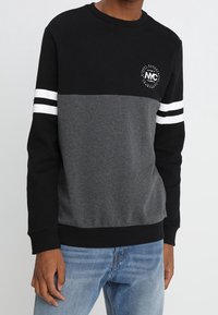 Homme portant un sweat-shirt à blocs de couleurs noir et gris avec des rayures blanches sur les manches et un petit logo circulaire sur la poitrine, associé à un jean bleu.