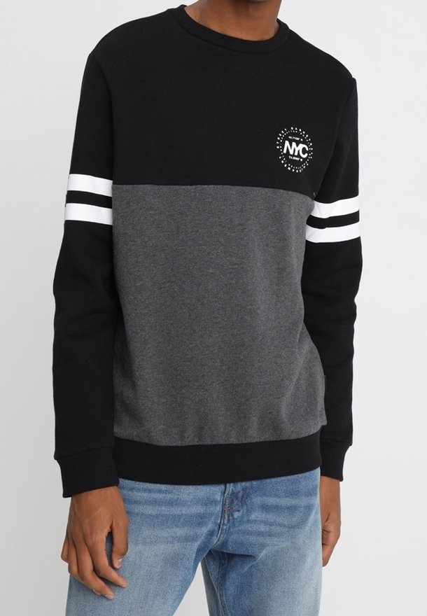 Homme portant un sweat-shirt à blocs de couleurs noir et gris avec des rayures blanches sur les manches et un petit logo circulaire sur la poitrine, associé à un jean bleu.