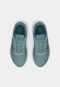 Zaļa Under Armour skriešanas apavu pāris ar baltām zoleēm un šņorēm, skatīts no augšas uz baltā fona.