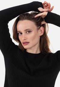 Pull noir côtelé à manches longues, doté d'un col rond et d'une texture douce. Le mannequin pose les mains dans les cheveux, sur un fond blanc.