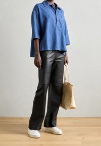 Oversized-Denimhemd mit Kragen, kombiniert mit schwarzen Lederhosen und weißen Sneakers. Eine beigefarbene Tragetasche ergänzt das Outfit.