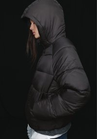 Žena u crnoj jakni s kapuljačom stoji u profilu s rukama u džepovima na tamnoj pozadini.