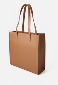 Ljusbrun rektangulär tygväska med långa handtag och texturerad yta, visad i en svag vinkel mot en enkel vit bakgrund.