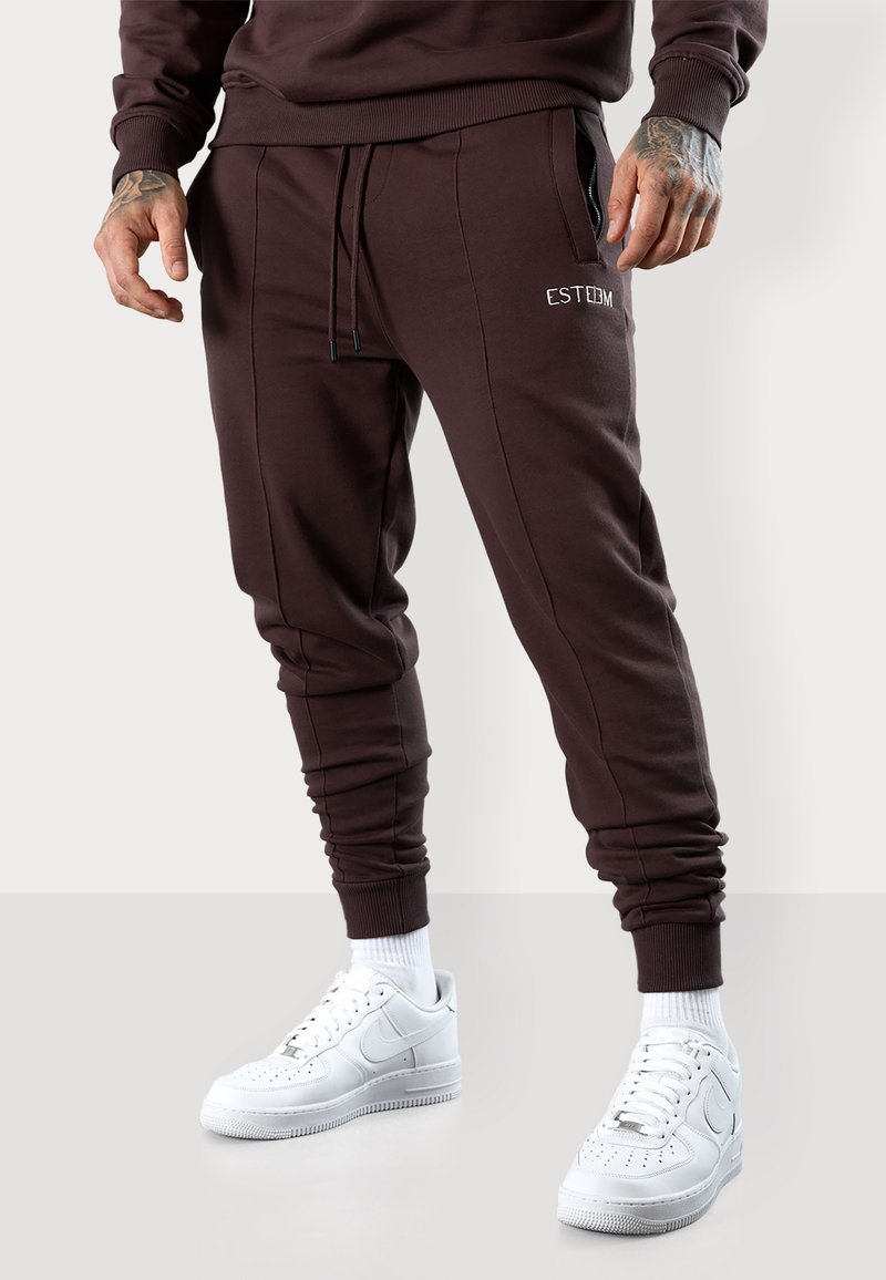 Dark brown jogger pants with elastic cuffs, featuring an embroidered logo and side pockets, paired with white athletic shoes and white socks.
