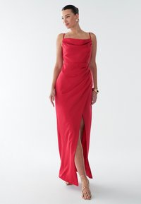 Červené saténové šaty s rolovaným výstrihom, tenkými ramienkami, záhybovým dizajnom a vysokým rozparkom. S doplnkami v podobe zlatých sandál s remienkami.