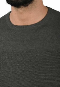 Pull en tricot vert foncé avec un col rond. La texture présente un motif tissé lisse et un design de point en relief contrastant sur la poitrine.