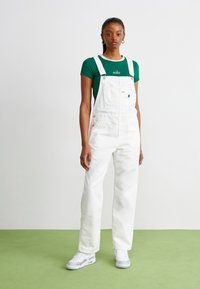 Salopette en denim blanc avec bretelles ajustables, poche avant et boutons latéraux. Assortie avec un t-shirt vert arborant un logo blanc. Baskets blanches.