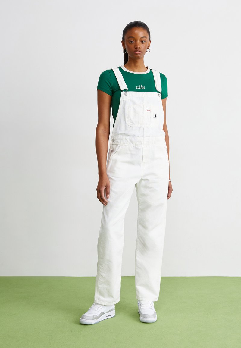 Salopette en denim blanc avec bretelles ajustables, poche avant et boutons latéraux. Assortie avec un t-shirt vert arborant un logo blanc. Baskets blanches.
