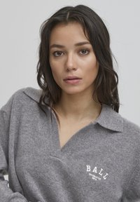 Suéter gris de punto con cuello tipo polo, con un corte en forma de V y texto blanco "BALL ORIGINATED 1974". Textura suave y corte relajado.