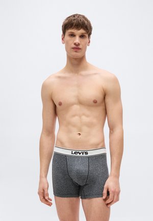 Νεαρός άνδρας στέκεται γυμνόστηθος, φορώντας γκρι μποξεράκια Levi's με λευκή μέση, ανοιχτόχρωμο φόντο.