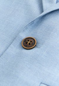 Svetlo modra tkanina z gladko teksturo, na kateri je rjav gumb z vklesano napisom "AUTHENTIC WEST DUDE". Preprost dizajn z drobno tkanino.