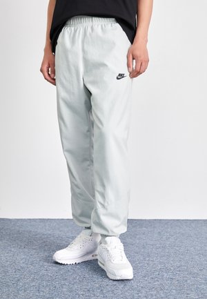 Person iført lysegrå Nike joggebukser og hvide Nike sneakers, stående på gråt tæppe foran en hvid væg.