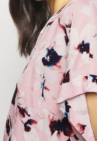 Femme portant un blouse rose avec de grands motifs floraux abstraits en marine, bordeaux et blanc, montrant une épaule et une partie du cou.