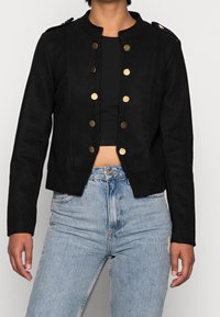 Giacca nera a bustino con bottoni dorati, caratterizzata da un colletto alla cinese e spalle strutturate, realizzata in un tessuto texturizzato. Design aderente.