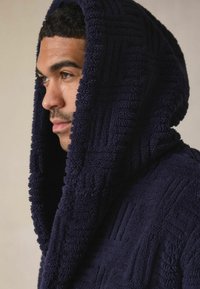 Granatowy szlafrok z kapturem wykonany z teksturowanego materiału o miękkiej, pluszowej powierzchni. Posiada geometryczny wzór oraz duży, głęboki kaptur.