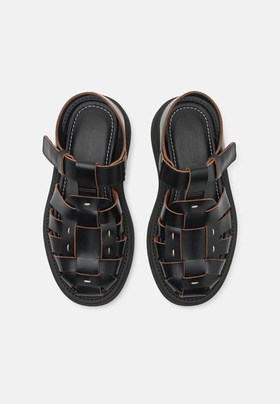 Sandales en cuir noir avec un design tressé, des coutures marron contrastantes, des côtés ouverts et une sangle pour la cheville sécurisée. Semelle texturée pour une meilleure accroche.