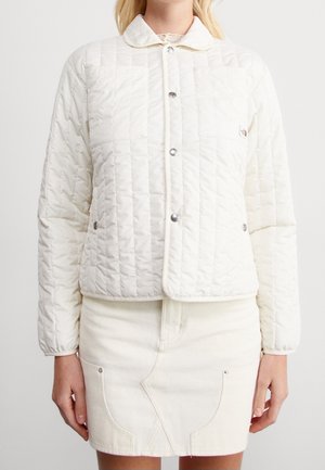 Femme portant une veste matelassée blanche avec des boutons-pression et une poche avant, associée à une jupe en denim blanche.