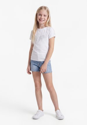 Jeune fille debout, souriante, portant un t-shirt blanc avec un texte, un short en jean bleu et des baskets blanches, sur un fond blanc uni.