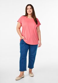 T-shirt color corallo a maniche corte con una vestibilità comoda e spacchi laterali, abbinato a pantaloni capri in denim blu. Il modello indossa delle ballerine beige chiaro.