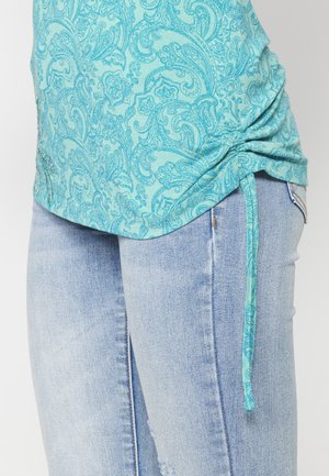 Turkio spalvos palaidinė su paisley raštu, susiraukšlėtomis šoninėmis dalimis ir reguliuojamais raišteliais, derinama su šviesiai plačiais džinsais, kurie turi aplamdytus akcentus.