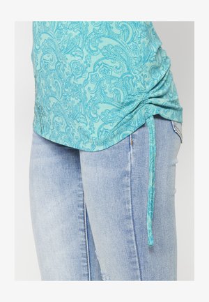 Türkiz színű, paisley mintás felső, ráncolt oldalakkal és állítható megkötőkkel, világos mosott farmerrel párosítva, amely sérült részletekkel rendelkezik.