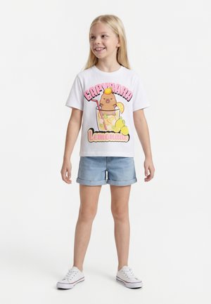 Jeune fille souriante portant un t-shirt blanc avec un capybara en dessin animé dans un verre de limonade et un short en jean, debout sur fond blanc.