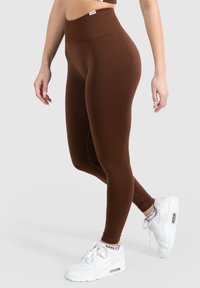 Hnědé legíny vysokého pasu vyrobené z elastického materiálu, s hladkou texturou a přiléhavým střihem, doplněné bílými teniskami.