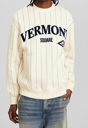 Osoba ubrana w kremowy sweter z cienkimi czarnymi pionowymi paskami i napisem "VERMONT SQUARE", zestawiony z niebieskimi dżinsami.