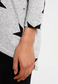 Pull gris avec motif d'étoiles noires ; présente un tissu tricoté texturé et des manches trois-quarts. Vue rapprochée d'une main reposant sur la manche.