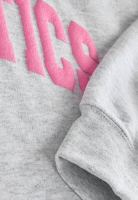 Grå sweatshirt med rosa filtbokstäver. Tyget har en mjuk textur, med synliga ribbade ärmslut och en avslappnad passform.