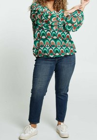 Femme portant un chemisier à motifs verts, marron et blancs, un jean bleu foncé et des baskets blanches, ajustant sa manche.