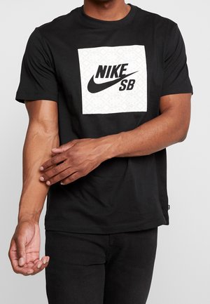 Homme portant un T-shirt noir Nike SB avec un logo carré blanc, ajustant sa manche, debout devant un fond clair uni.