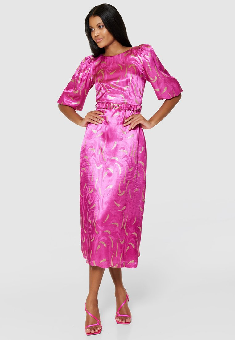 Abito di raso rosa con maniche a balzo, vita con cintura e motivi astratti dorati. Lunghezza alla caviglia, abbinato a sandali rosa con cinturini.