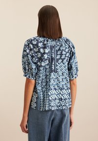 Blusa blu navy con stampa floreale, maniche a palloncino, scollo arricciato e vestibilità morbida, abbinata a pantaloni di jeans azzurri.