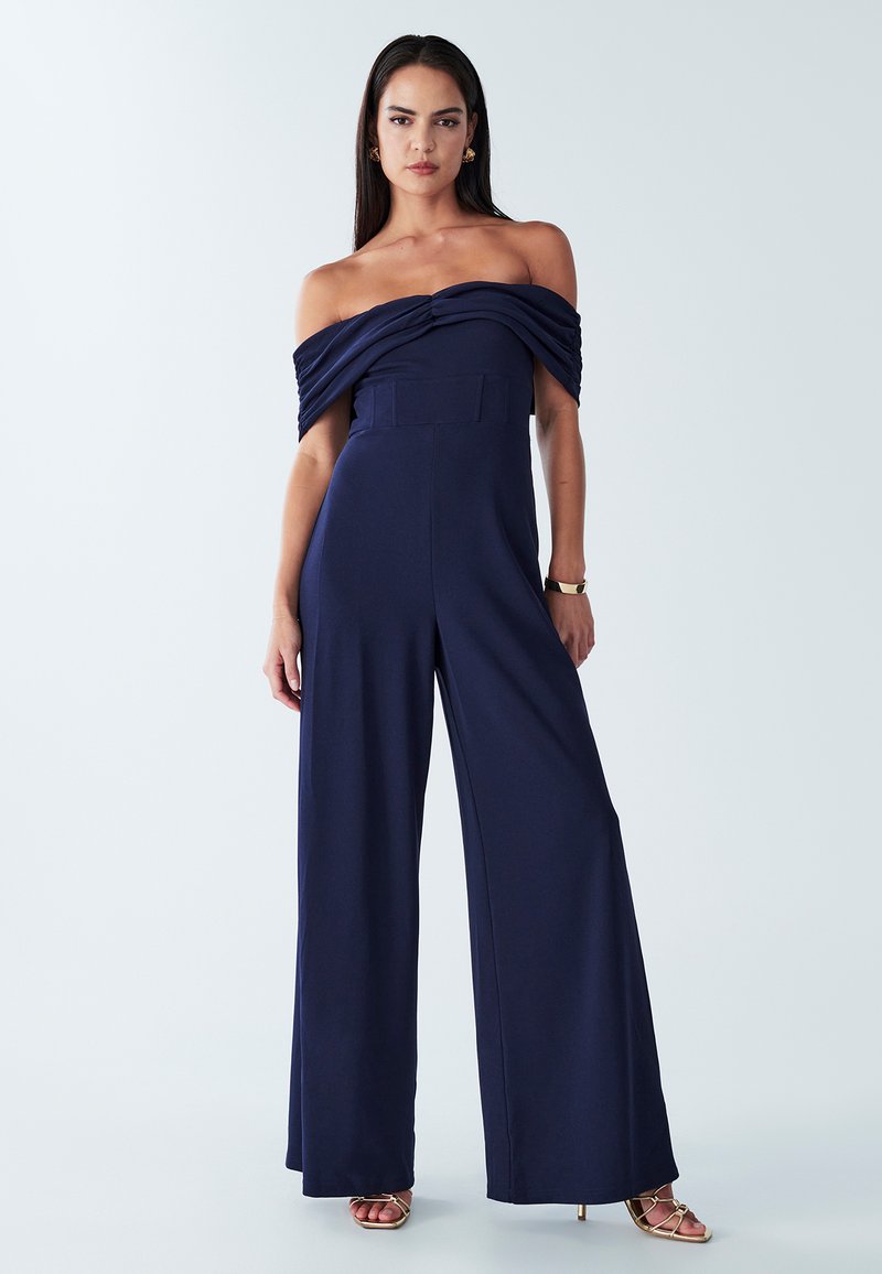 Marineblauwe off-the-shoulder jumpsuit met gedrapeerde details, wijde pijpen en een getailleerde top. Glad stof met minimale hardware.