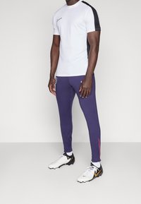 Weißes Kurzarm-Sportshirt mit schwarzem Seitenpanel; lila enganliegende Hose mit roten Akzenten; schwarz-weiße Schuhe mit orangefarbenen Details.