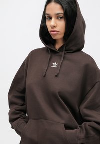 Καφέ φούτερ με κουκούλα από μαλακό ύφασμα, με τσέπη μπροστά, κορδόνι για τη ρύθμιση της κουκούλας και μικρό λευκό λογότυπο adidas στο στήθος.