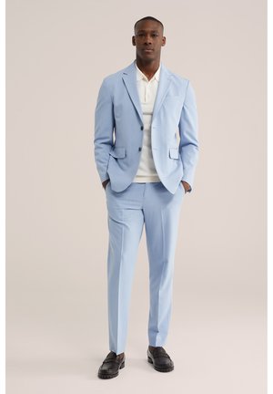 Man draagt lichtblauw pak met wit overhemd en zwarte loafers, staand met handen in de zakken tegen een effen achtergrond.