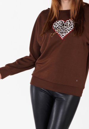Kobieta w brązowej bluzce z długim rękawem, ozdobionej sercem w panterkę i złotym napisem, w połączeniu z czarnymi błyszczącymi legginsami z ekologicznej skóry.